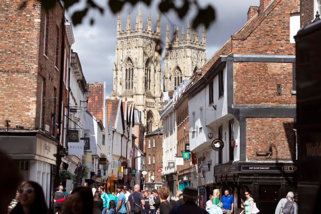 Bustling street in York with historic buildings, shoppers, and the towering York Minster in the background under a cloudy sky.