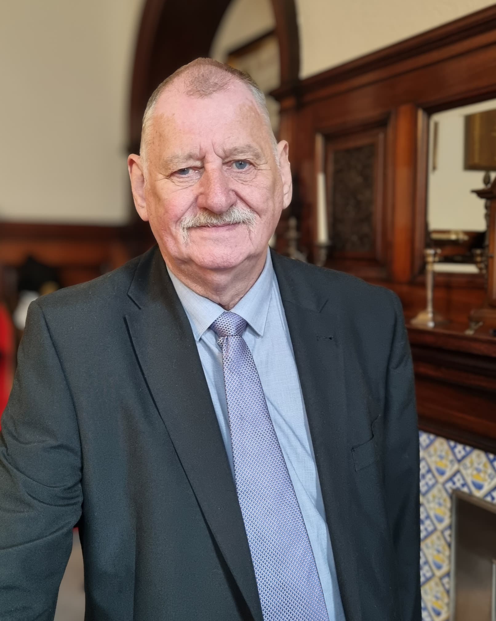 Elderly man in a suit standing by a wooden fireplace