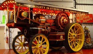 Vintage steam-powered fairground organ with intricate designs and bright lights in a museum setting.
