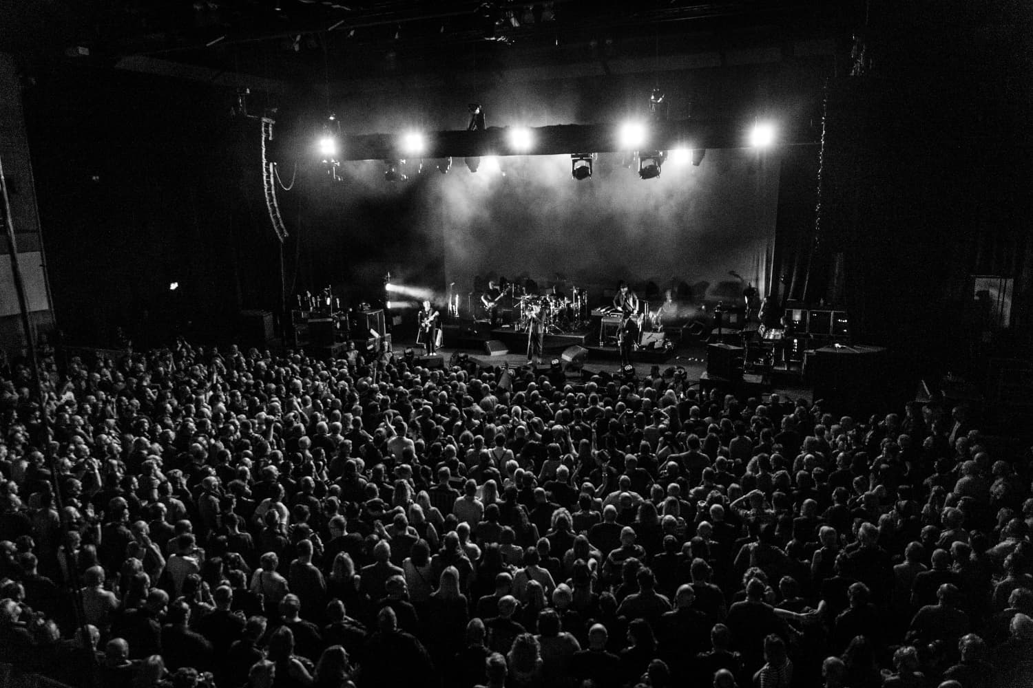 Crowded concert hall with band performing on stage under bright lights, audience seated and standing in darkened space.