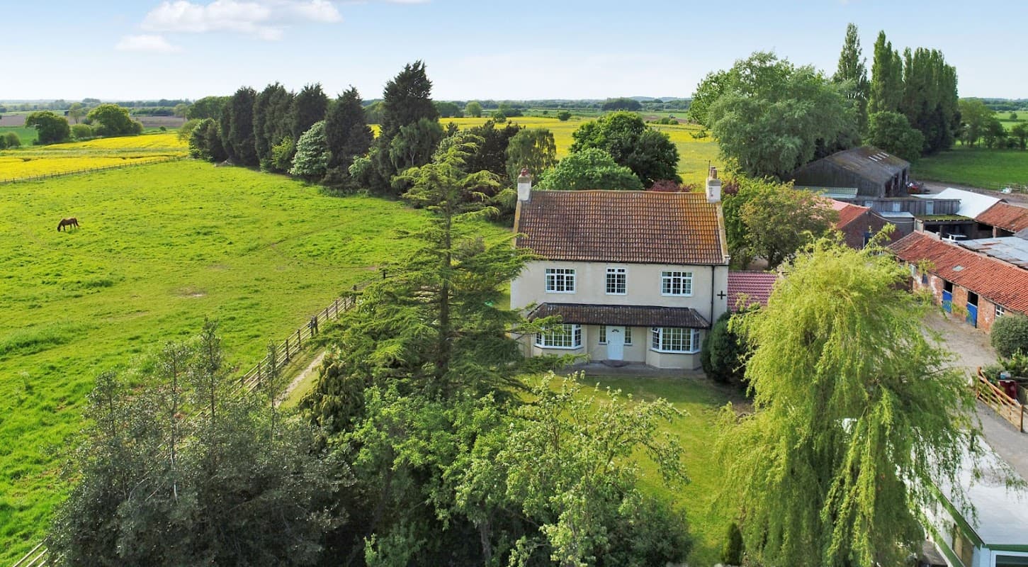 Aerial view of a house surrounded by greenery, fields, and trees, with horses grazing in the distance.