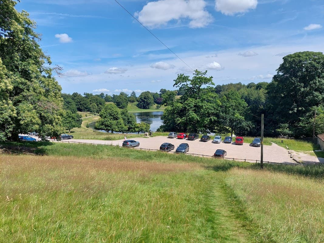 Free parking area with cars, surrounded by lush greenery and a pond under a blue sky with scattered clouds.
