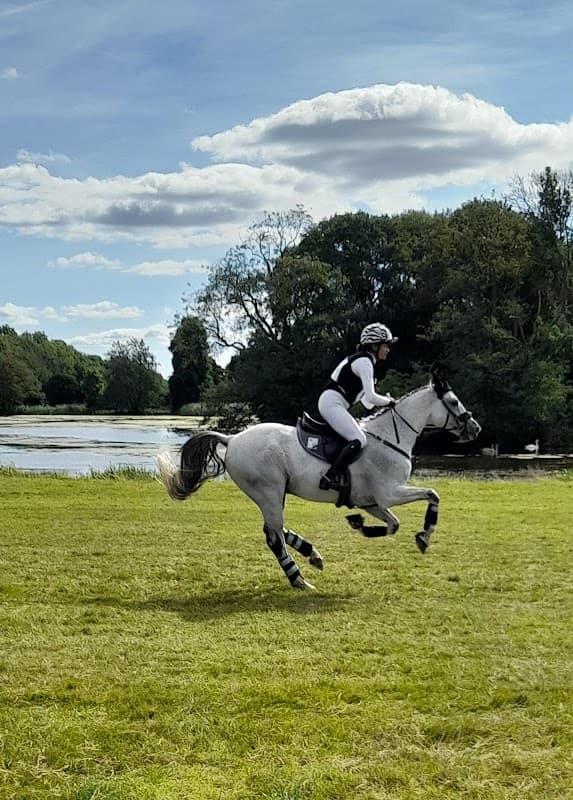 A rider in white attire gallops on a gray horse across a grassy field near a lake and trees under a blue sky.