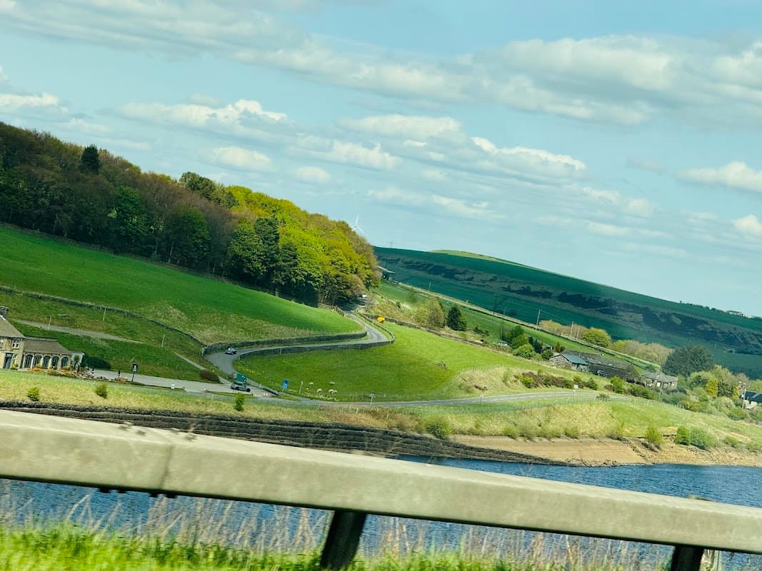 Lush green hills and winding roads near Barkisland Park, with a scenic view of water and distant fields under a blue sky.