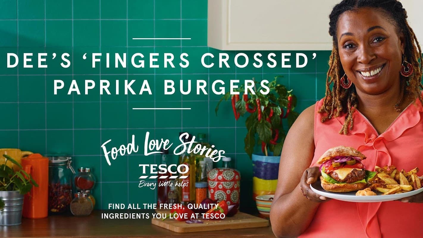 A woman smiles while holding a plate of paprika burgers and fries in a vibrant kitchen with green tiles.