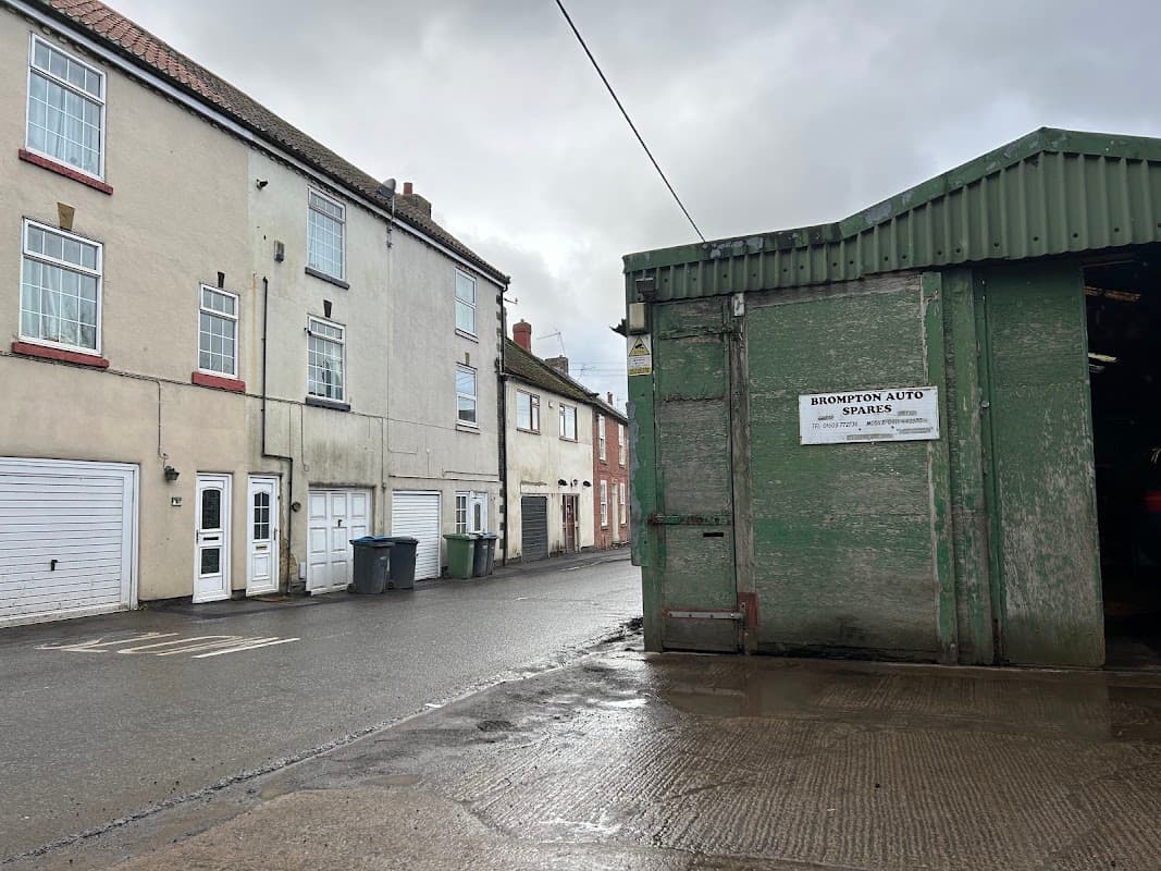 Brompton Auto Spares sign on a green building beside residential houses and parked bins on a wet street.