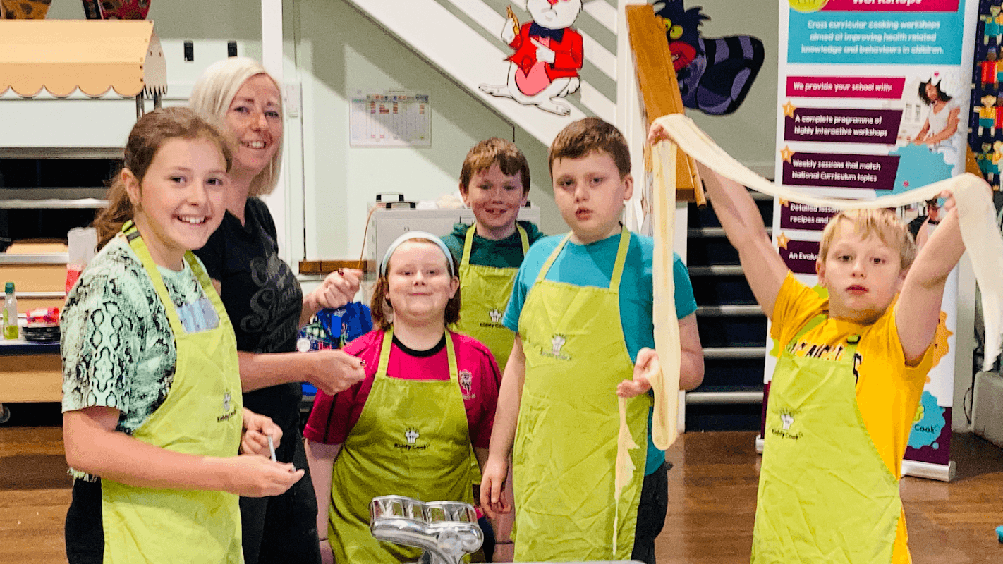 Children in green aprons cooking with an instructor, preparing food in a bright, cheerful kitchen setting.