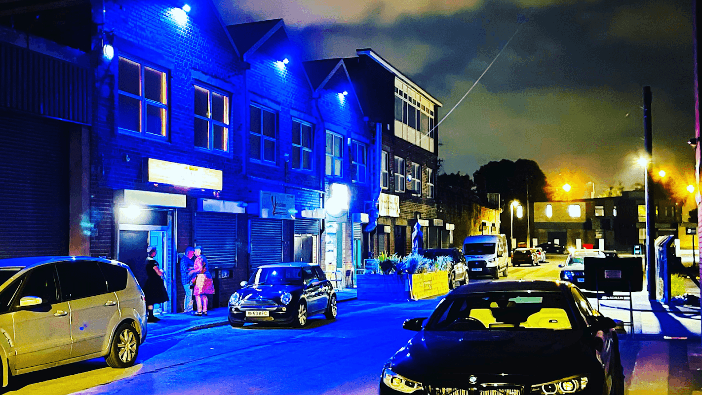 Nighttime scene of Tapestry Arts bar with blue lights, parked cars, and people outside in Dudley Hill, Yorkshire.