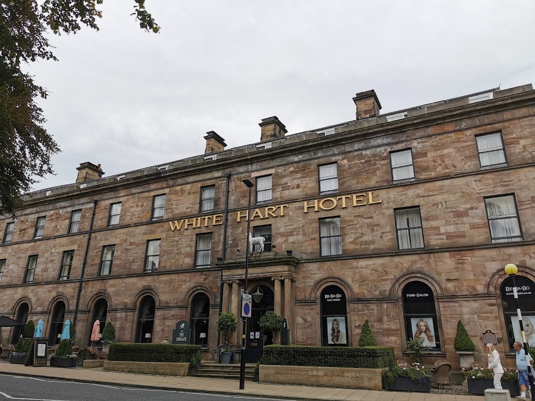 Historic stone building with large windows, "WHITE HART HOTEL" sign, and decorative plants in front.