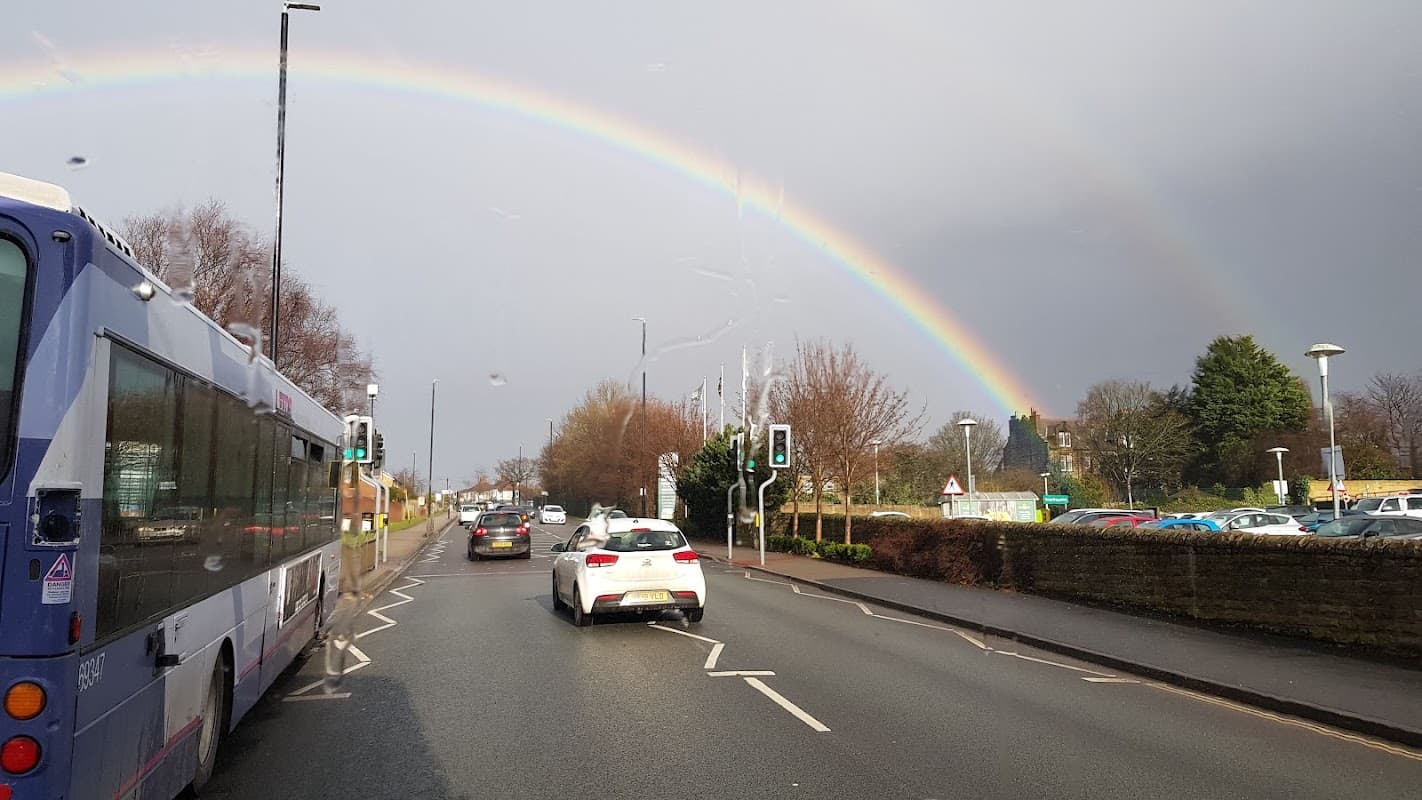 A bus and cars on a road with a vibrant rainbow arching over trees and buildings in Horsforth, Yorkshire.