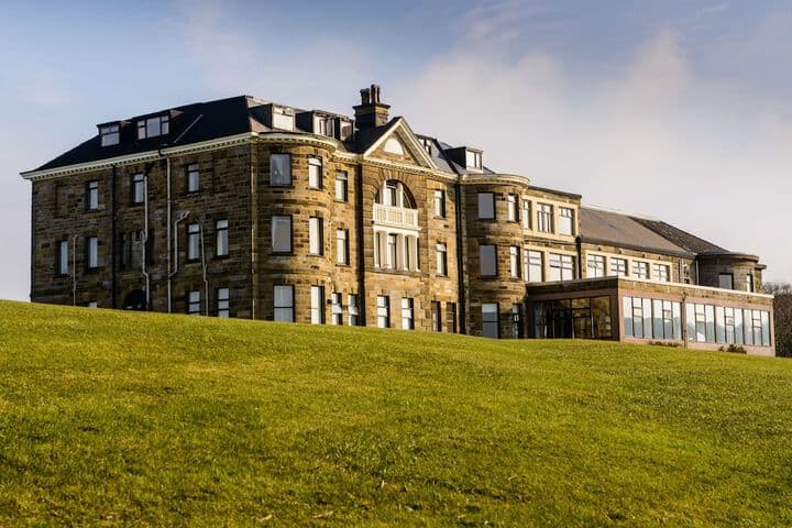 Historic stone mansion with large windows, surrounded by lush green lawn and a glass-enclosed extension.