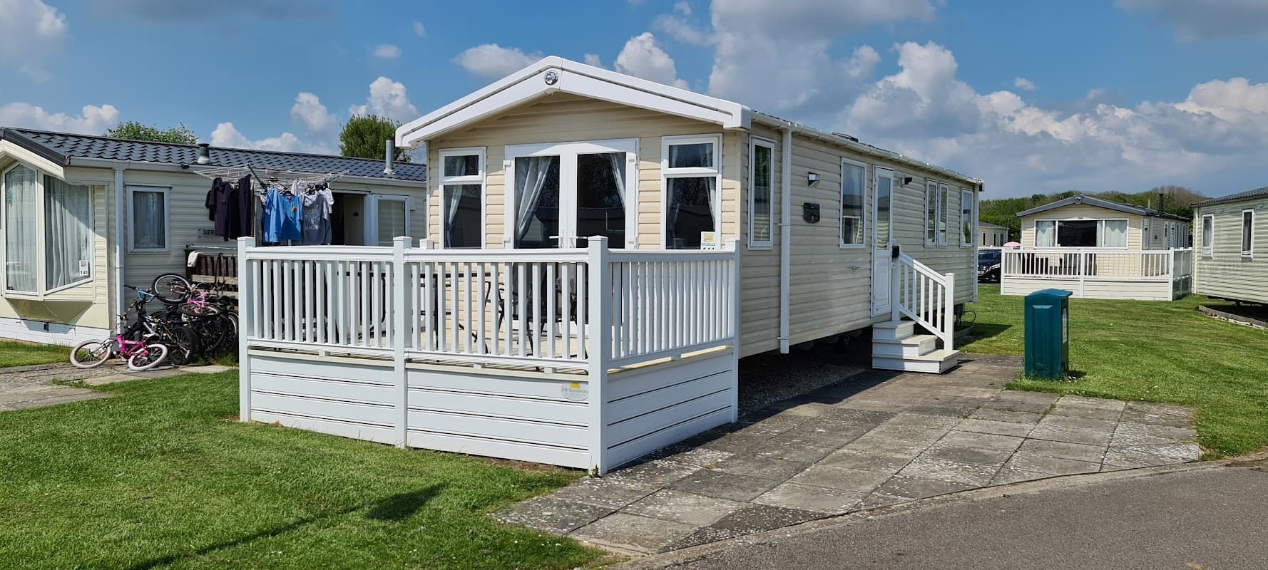 Wooden caravan with a deck, surrounded by green grass and nearby caravans, under a blue sky with fluffy clouds.