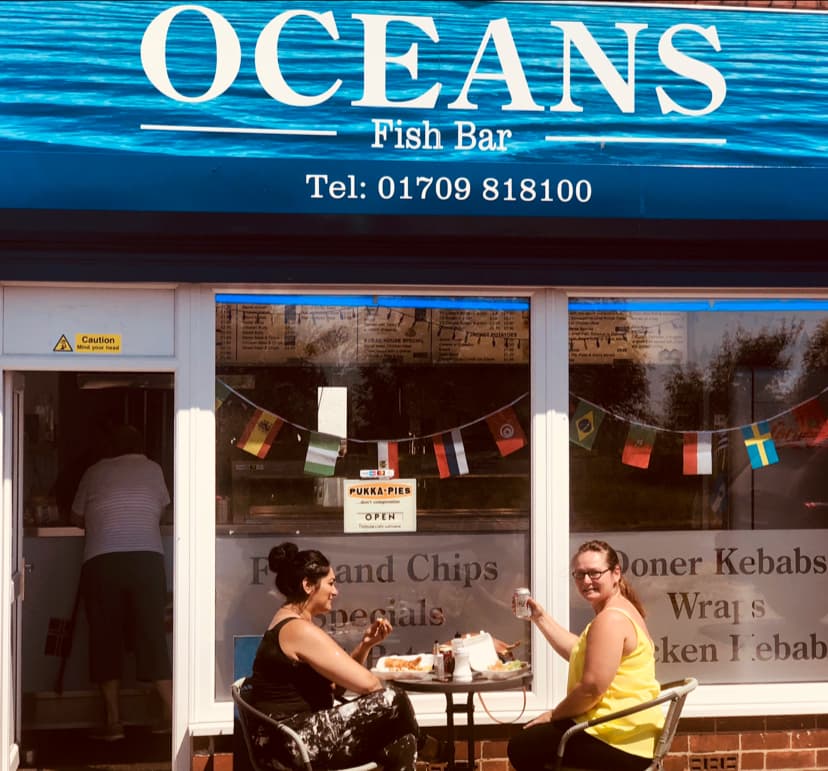 Sign for Oceans Fish Bar with flags, two women dining outside, enjoying food and drinks in Maltby, Yorkshire.