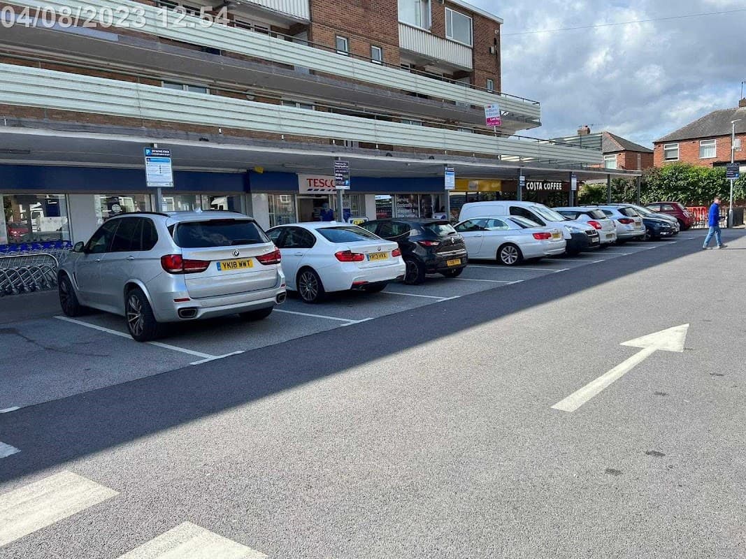 A row of parked cars in a car park with a Tesco store in the background and a cloudy sky above.
