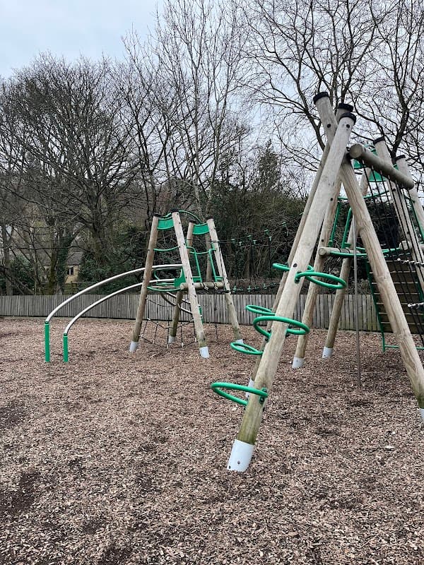 Playground equipment featuring climbing frames, slides, and green accents, surrounded by trees and wood chip ground.