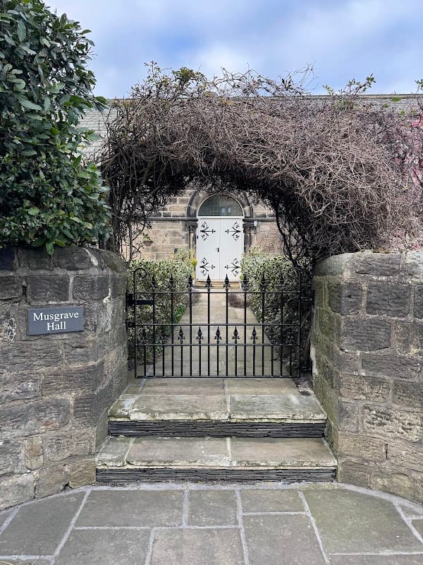 Archway covered in vines leads to a white door at Musgrave Hall, framed by stone walls and a paved pathway.
