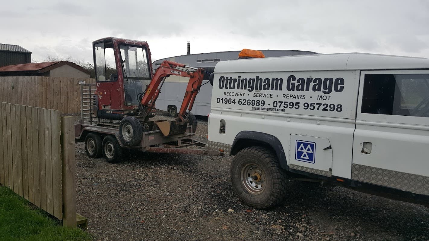 Red mini excavator on a trailer next to a service vehicle with "Ottringham Garage" branding. Gravel surface and cloudy sky.