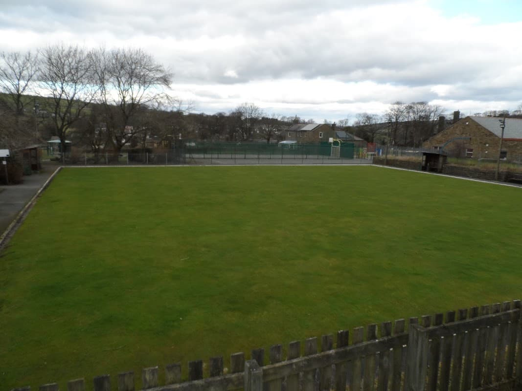 Lush green bowling green surrounded by trees and buildings under a cloudy sky in Oxenhope, Yorkshire.