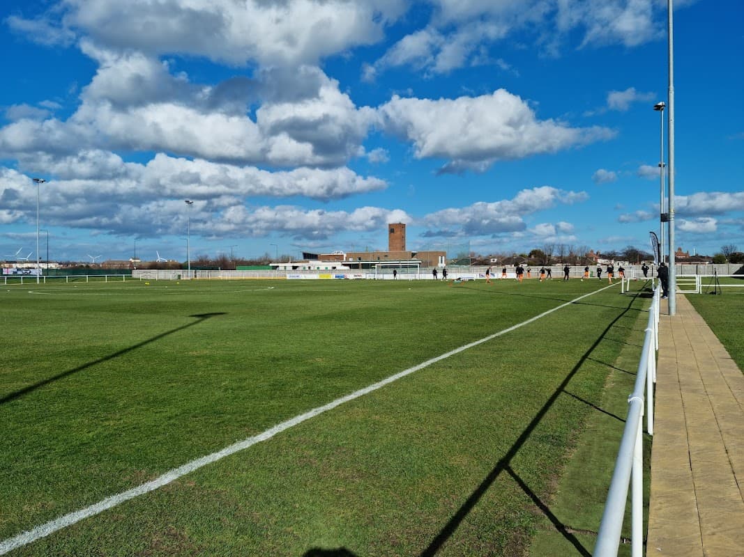 Green football pitch with white lines, a blue sky, and clouds; background features a building and a tower.