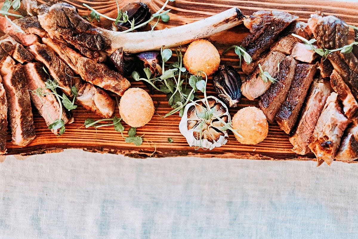 A wooden platter featuring an assortment of grilled meats, garnished with herbs and round orange croquettes.