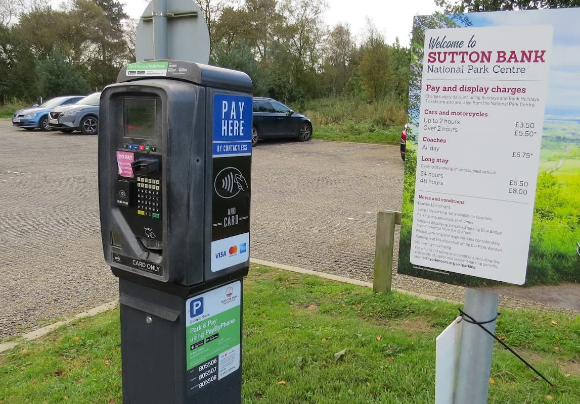 Pay & Display machine and sign detailing parking charges at Sutton Bank Car Park, surrounded by greenery and vehicles.