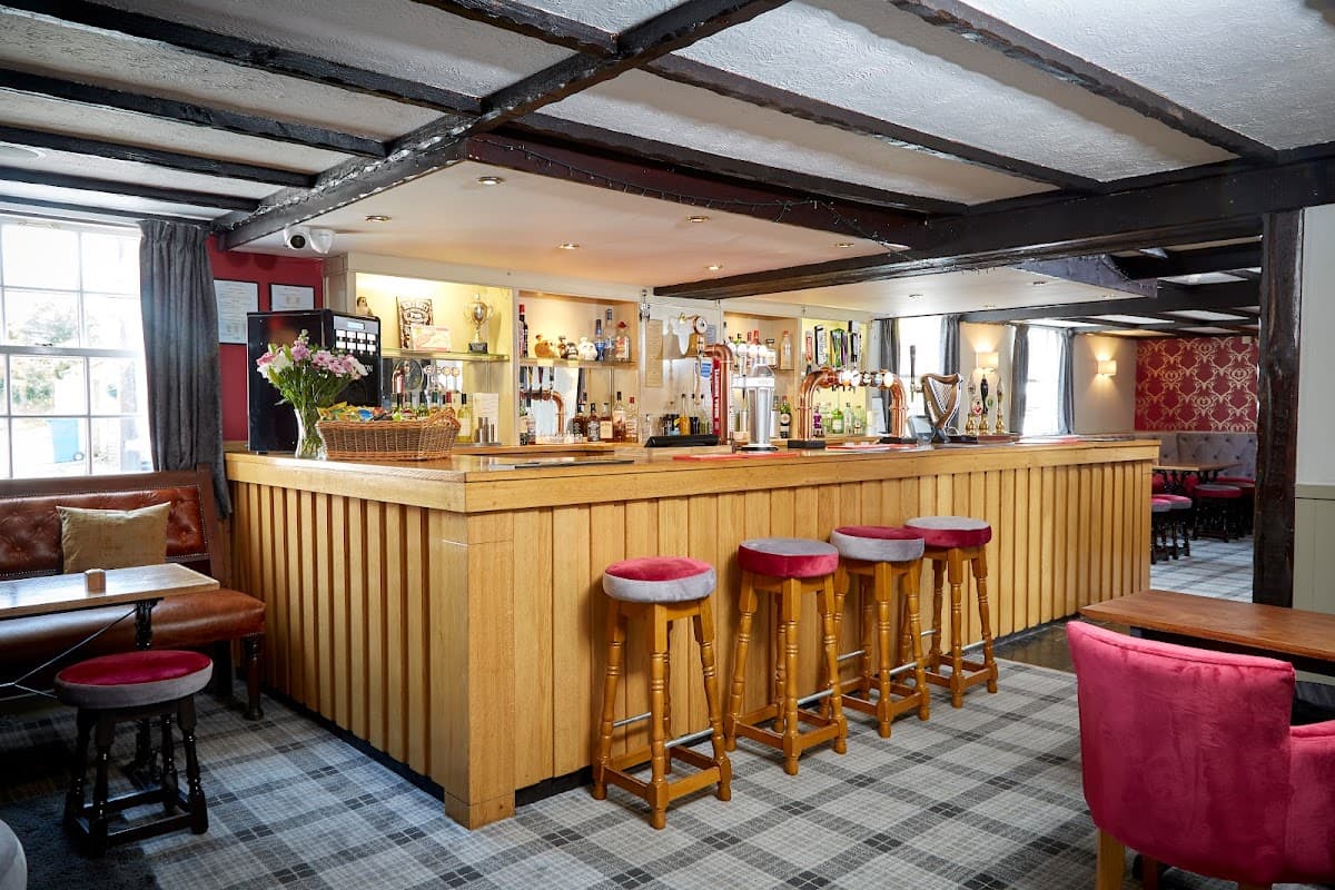 Warm wooden bar with stools, drinks on display, and cozy seating in a charming pub interior with patterned wallpaper.