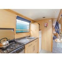 "Andromeda narrowboat interior with wooden cabinets, stainless steel sink, black stove, and cozy seating area."