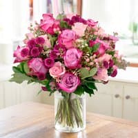 Pink and purple rose bouquet with greenery in a clear glass vase on a wooden table.