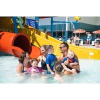Family enjoying water park with red and yellow slides, surrounded by sun umbrellas and other visitors.