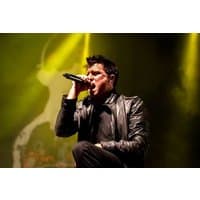 Black leather jacket with microphone, singer performing energetically on stage with yellow lighting in the background.