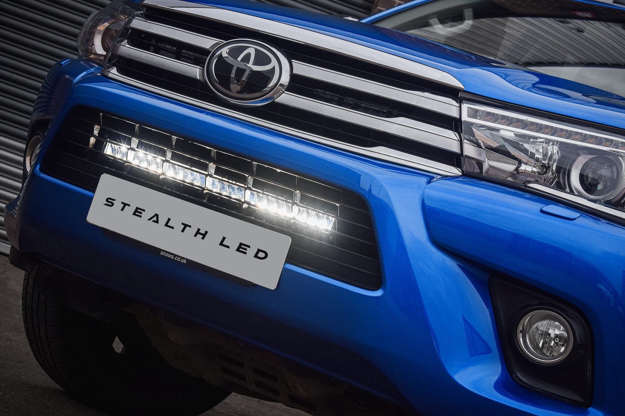 Blue Toyota Hilux MK8 with a sleek black grille featuring a luminous LED integration kit, showcasing bright white lights and a prominent "STEALTH LED" logo.