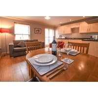 Dining table set for two in cozy apartment with sofa, wooden floors, modern kitchen, and warm lighting.
