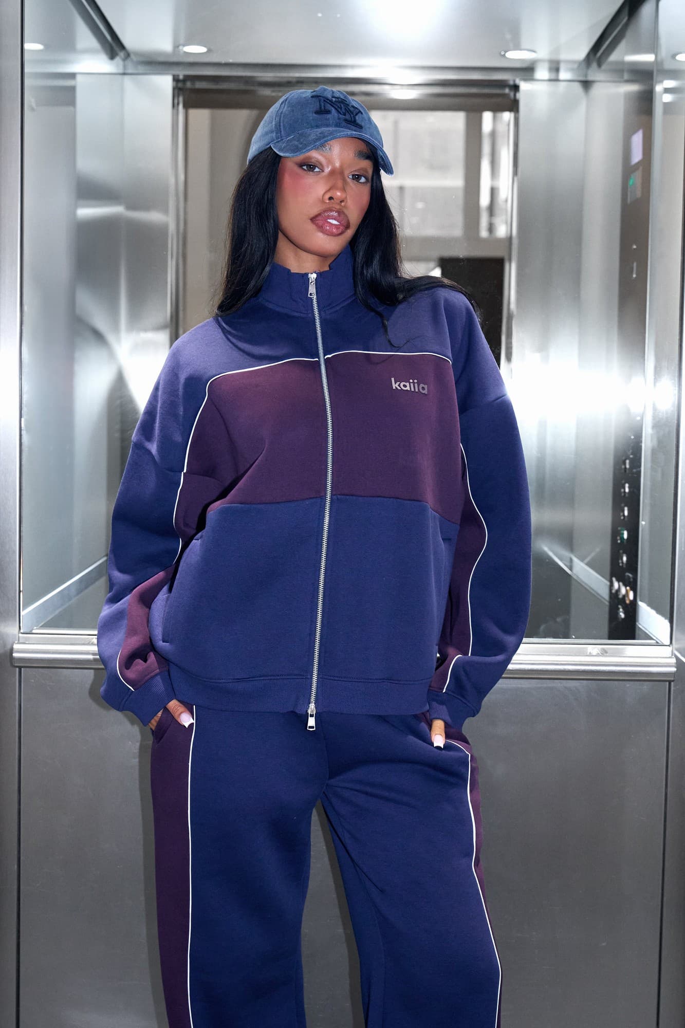 Oversized zip-through sweatshirt in navy and burgundy, featuring contrasting panels, a high collar, and side pockets, paired with matching pants and a navy cap.
