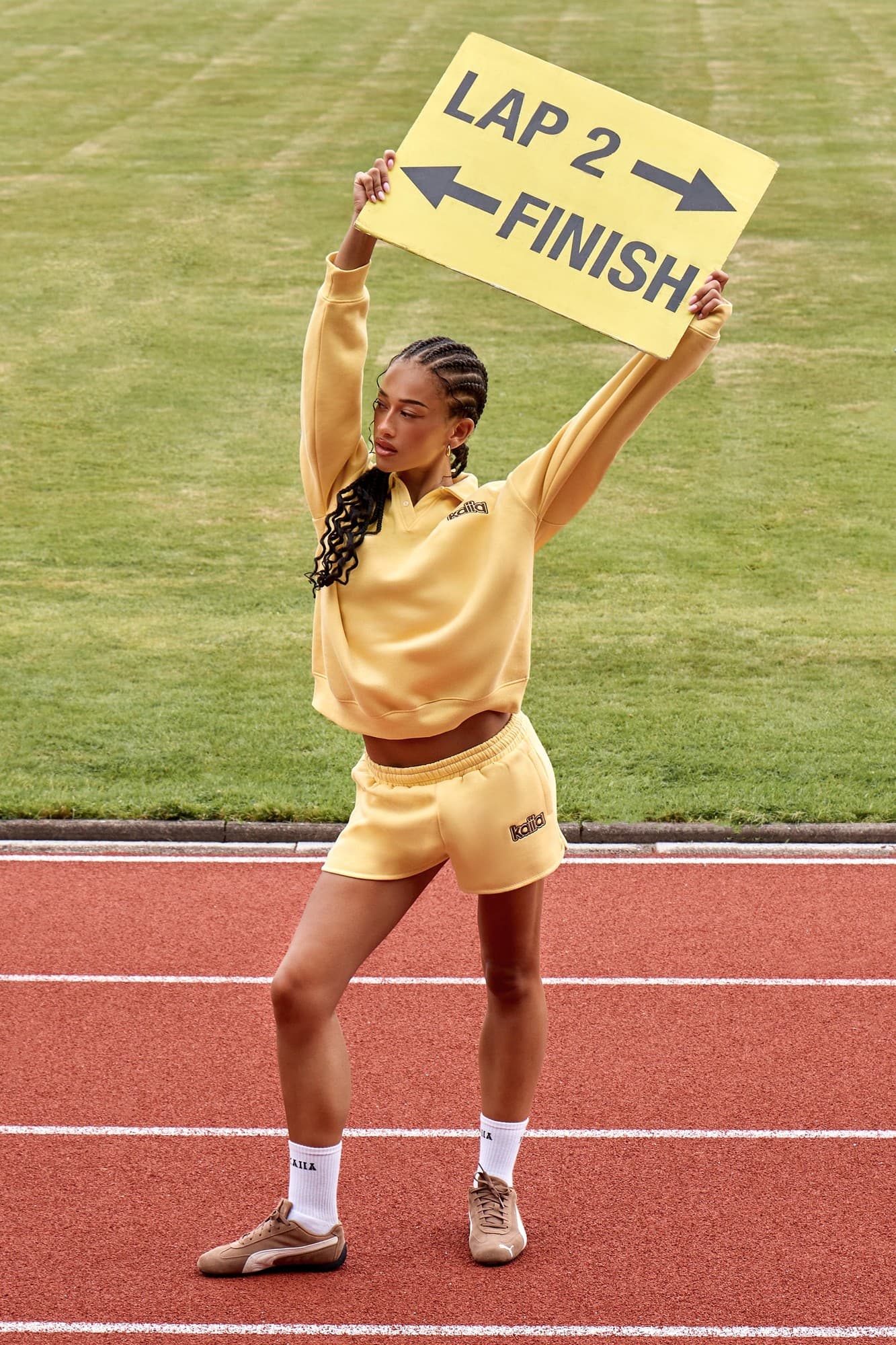 Yellow mini sweat shorts featuring the Kaiia logo, paired with a matching top, worn by a model holding a sign that says "LAP 2" and "FINISH," set on a running track.