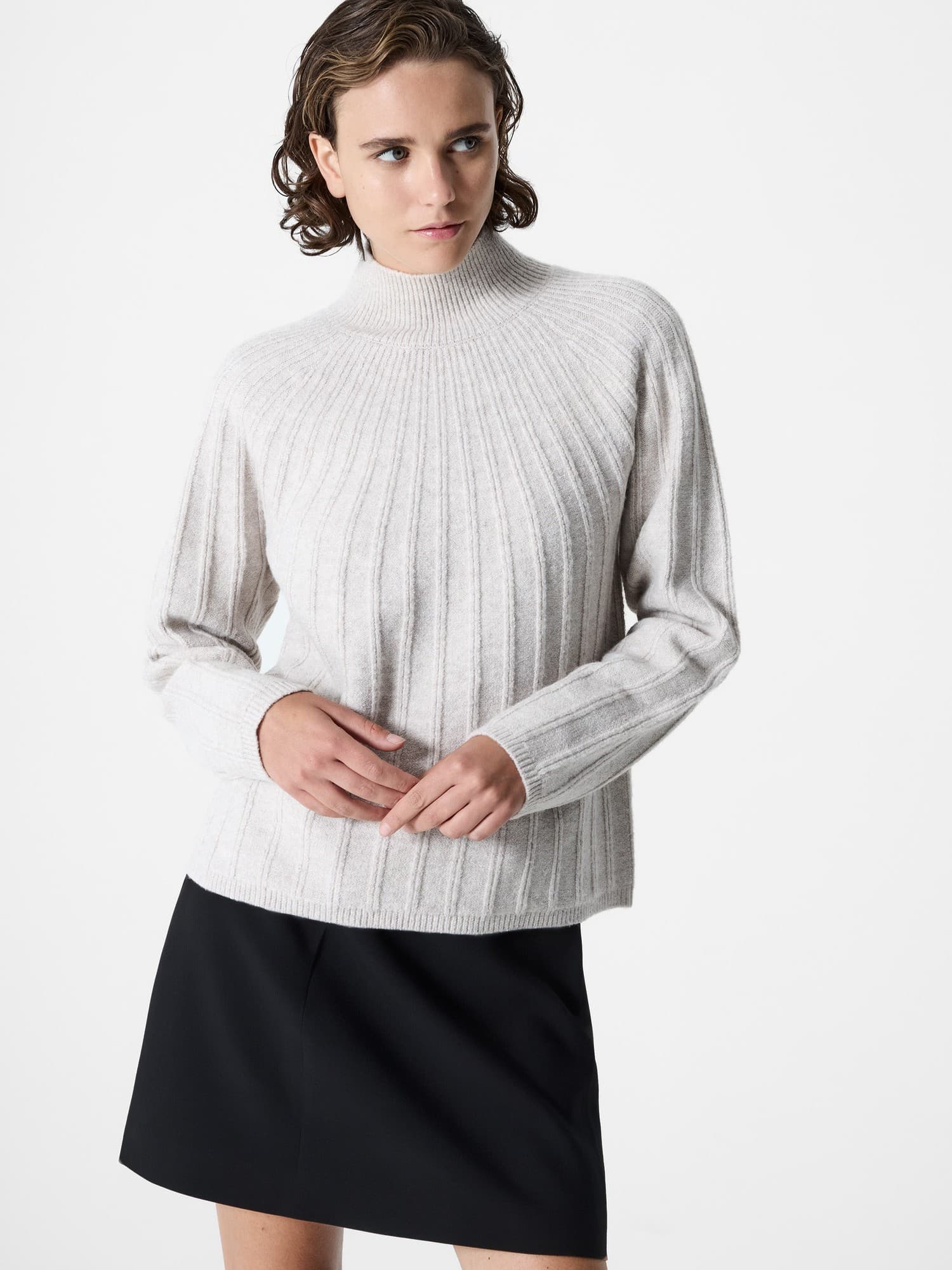 Light beige ribbed knit jumper with a high neck and long sleeves, featuring a relaxed fit, paired with a black skirt for a stylish look.