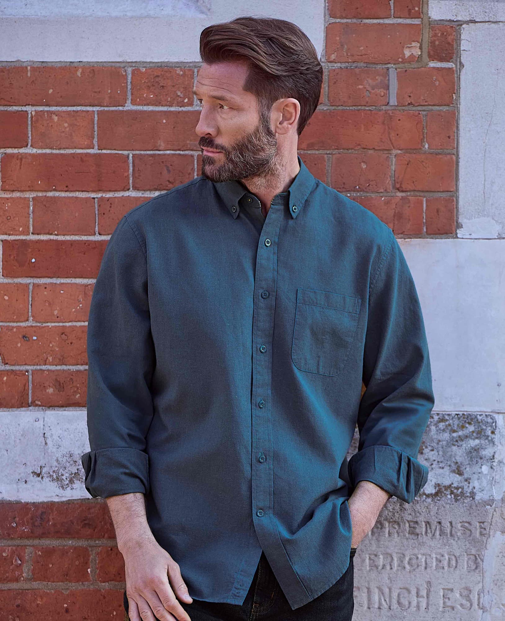 Dark blue classic fit button-down casual shirt made from a linen-cotton blend, featuring a chest pocket and rolled-up sleeves, worn by a man against a brick wall.