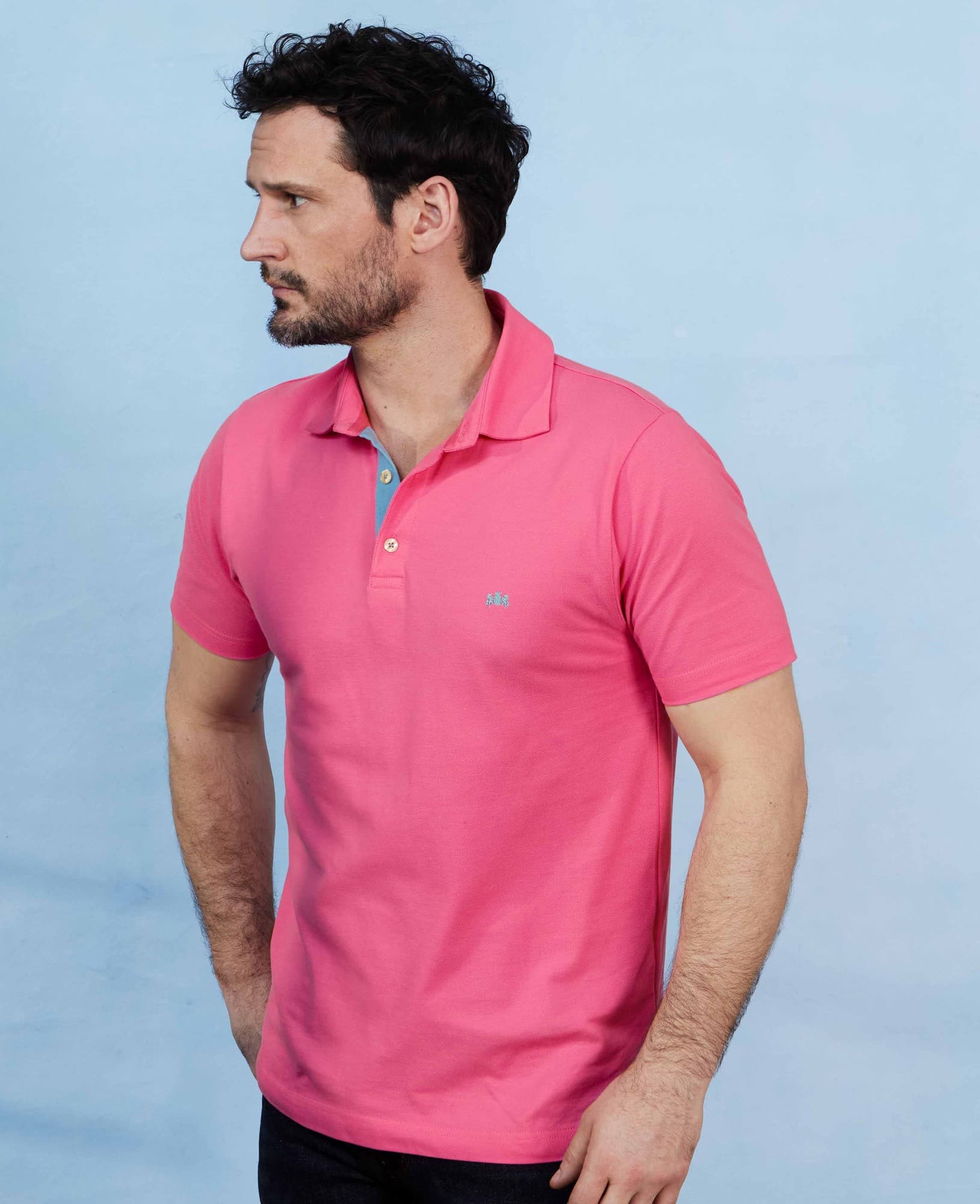 Men's short sleeve polo shirt in vibrant pink cotton pique, featuring a classic collar and a subtle logo on the chest, shown on a model against a light blue background.