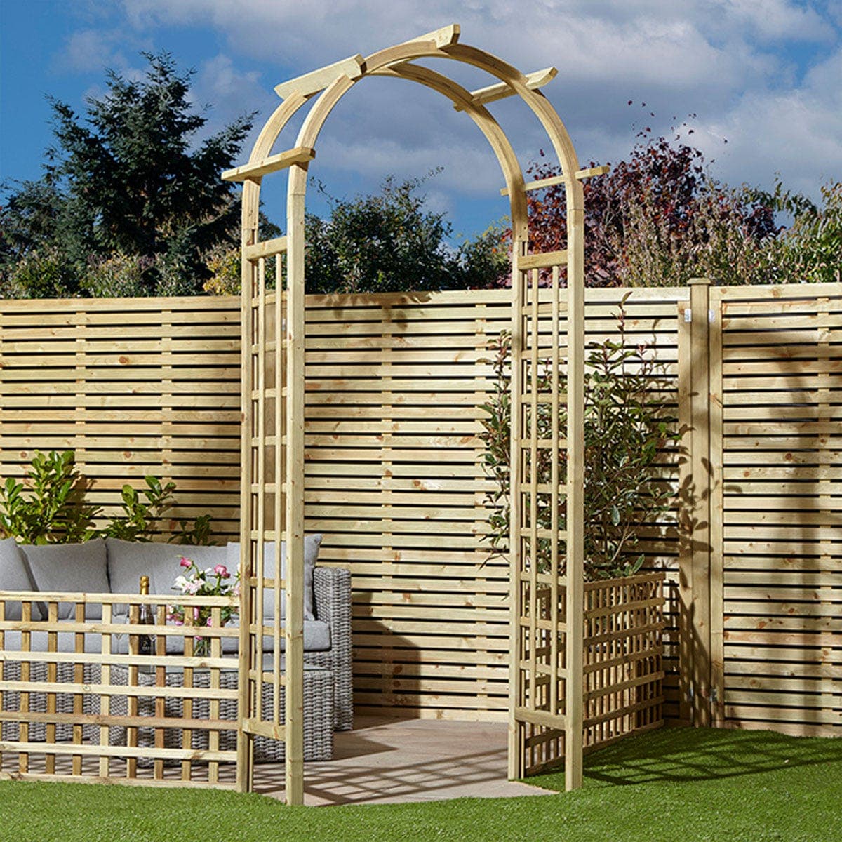 Natural pressure-treated timber arch with a curved top and trellis sides, set against a wooden fence, surrounded by greenery and outdoor furniture.