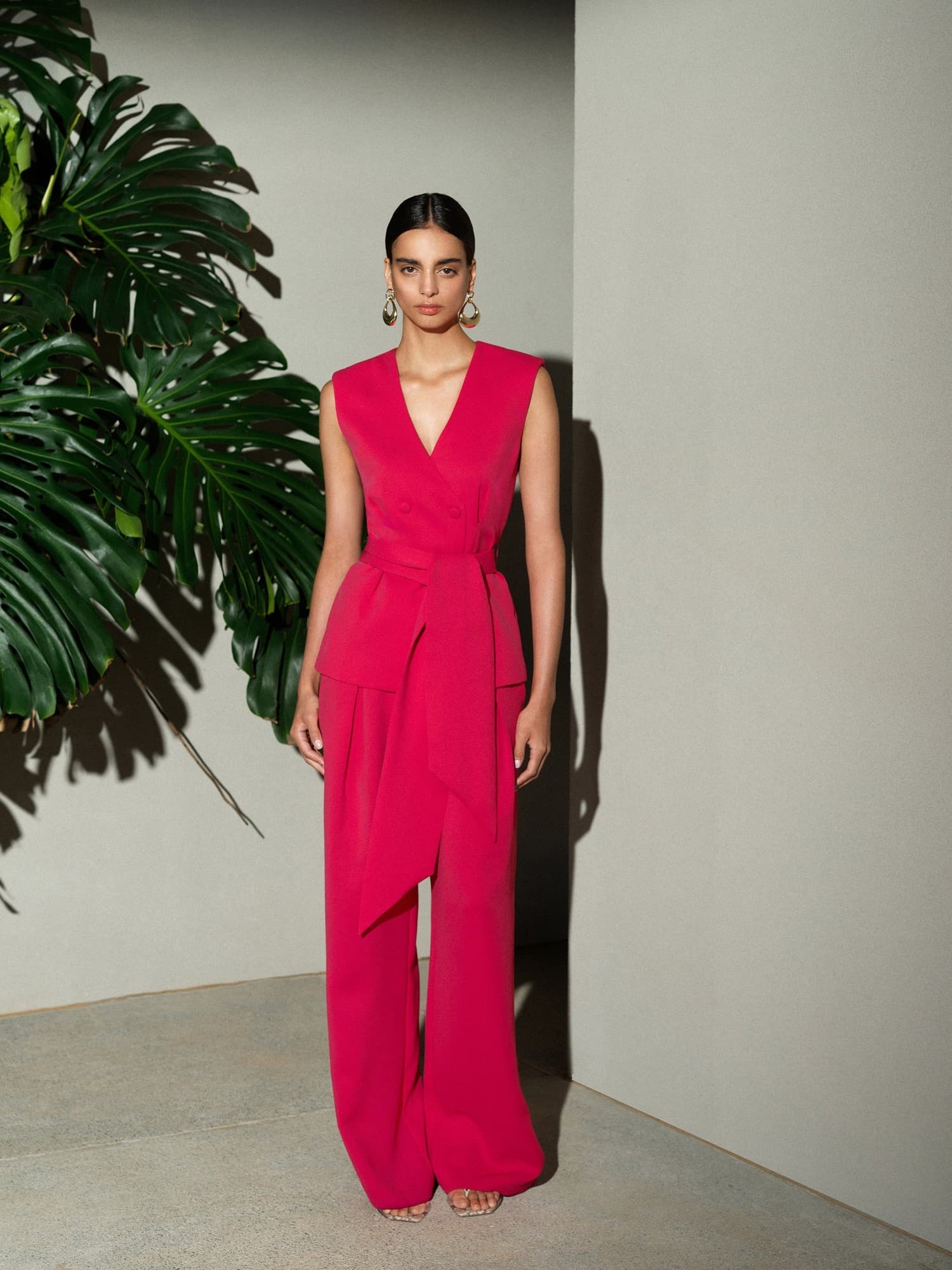 Peony pink self-tie waistcoat with a deep V-neck, featuring a tailored fit and wide leg trousers, styled against a minimalist backdrop with tropical foliage.