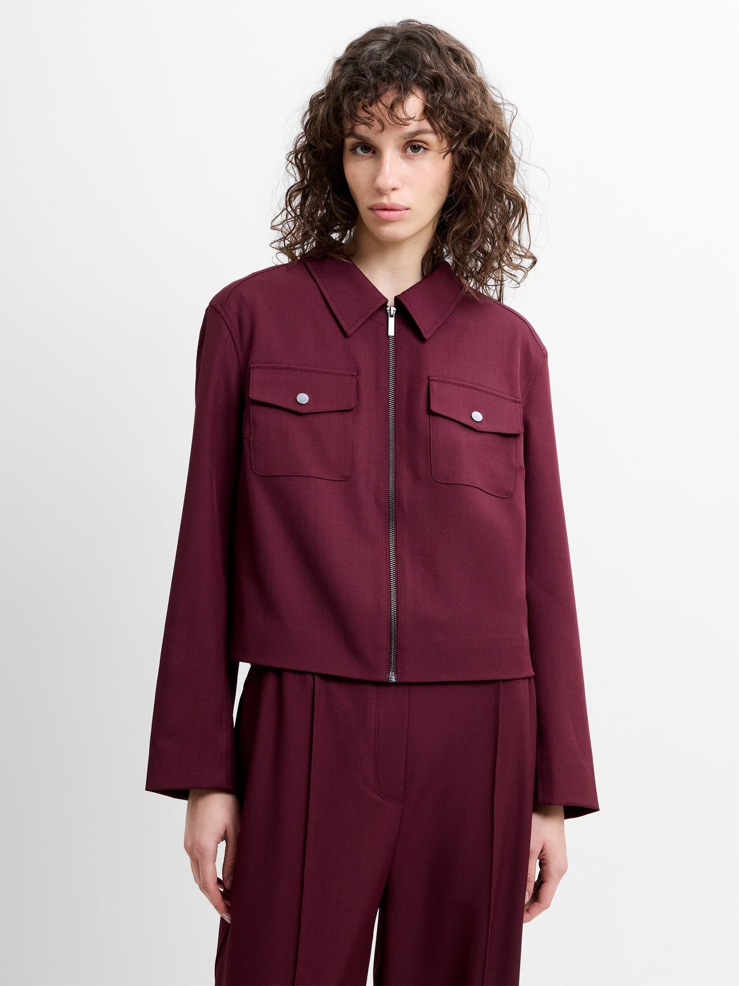 Structured hem jacket in deep burgundy, featuring a zip front, two chest pockets with silver buttons, and long sleeves, made from a tailored suiting material.