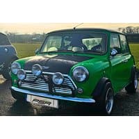 Green classic Mini car with black hood, chrome grille, and four front-mounted spotlights.