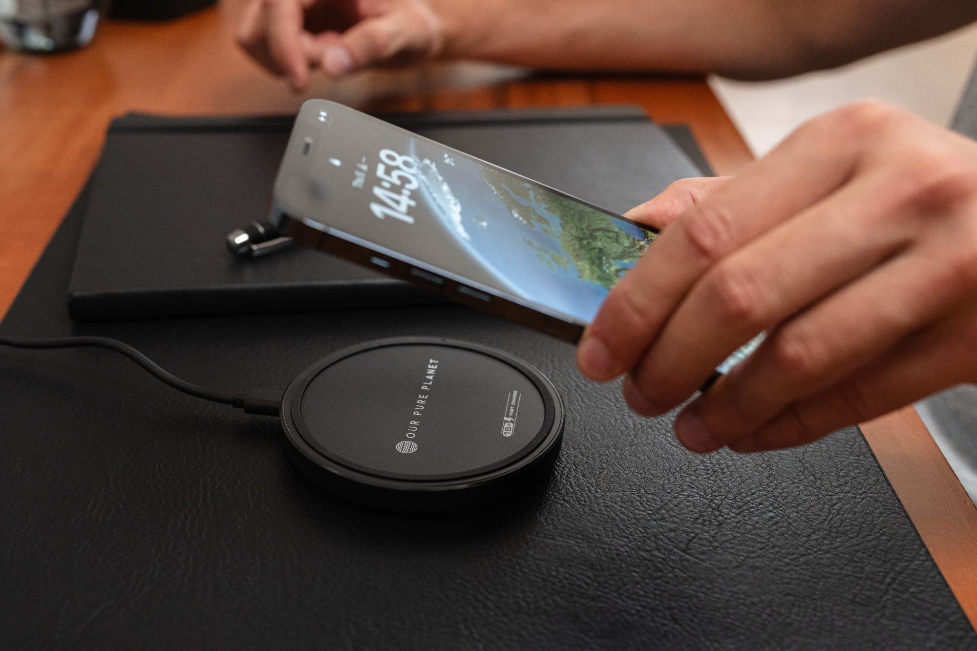 Black 15W wireless charging pad with a smooth surface, featuring the brand name "OUR PURE PLANET," as a hand holds a smartphone above it, ready to charge.