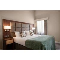 Hotel room with a large bed, green throw, two pillows, wooden headboard, bedside lamps, and a window with curtains.