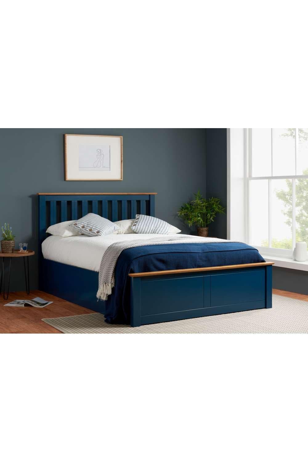 Solid wood king-size ottoman storage bed frame in navy with a gas lift mechanism, featuring a slatted headboard and a contrasting wood trim, set against a light wall and natural light.