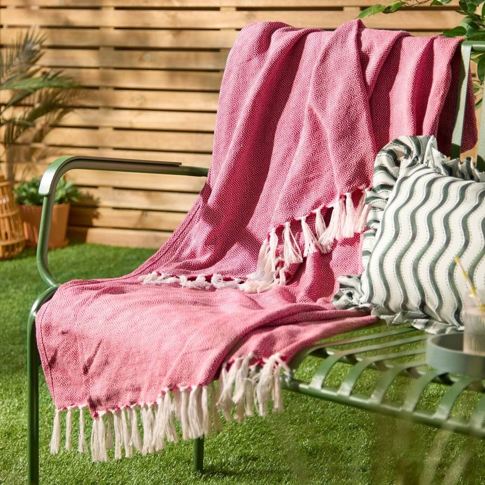 Woven pink throw blanket with white fringes draped over a green metal chair, complemented by a patterned cushion, set against a wooden backdrop and lush greenery.