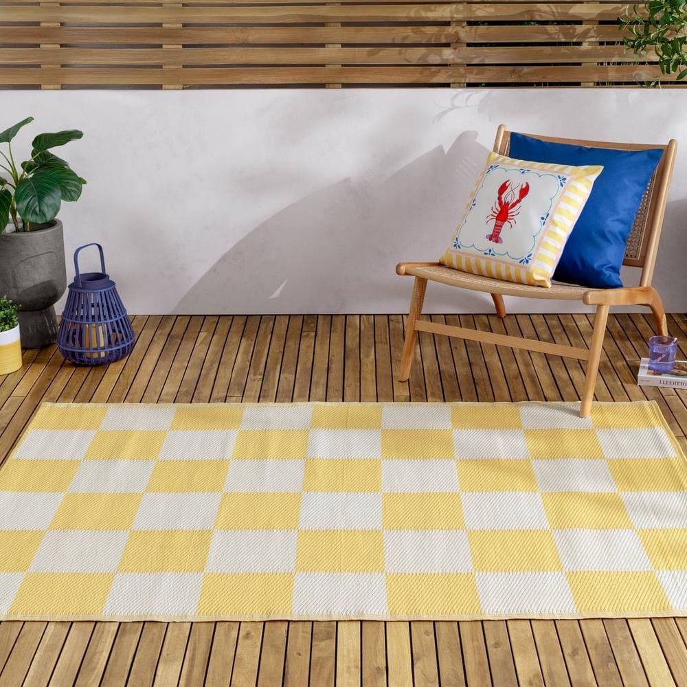 Yellow and white checkerboard outdoor rug made of durable material, placed on a wooden deck alongside a blue pillow, a potted plant, and a lantern.