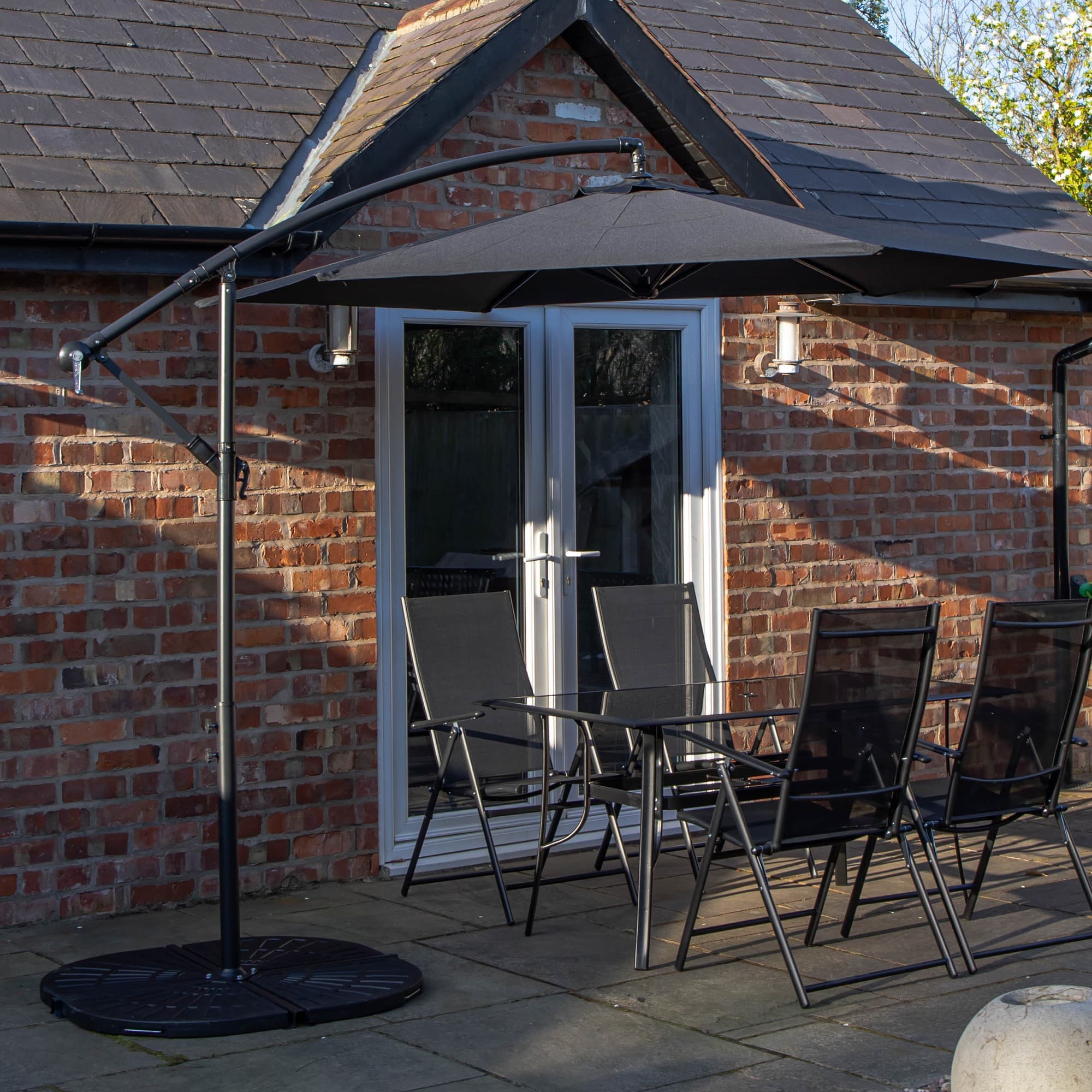 Rectangular glass top table with four black chairs, accompanied by a black banana parasol and sturdy base, set against a brick wall and patio.