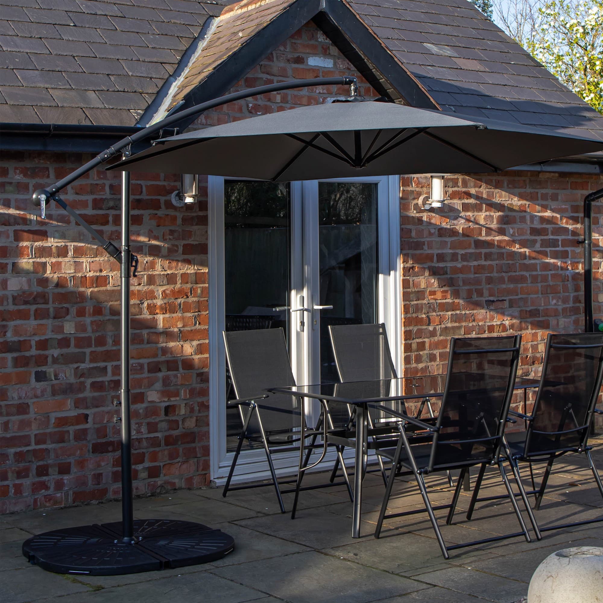 Rectangular glass top table with four black chairs under a grey banana parasol, set against a brick wall and patio.