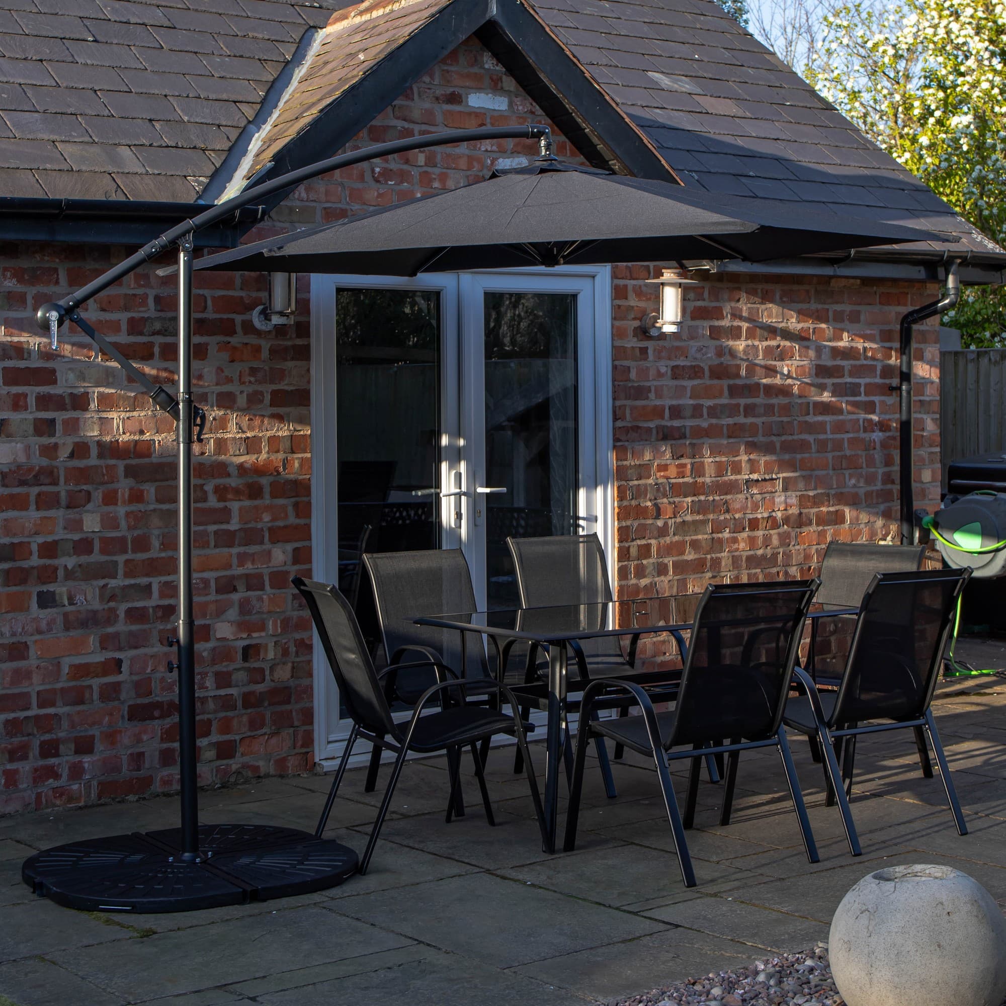 Rectangular glass-top dining table with six black chairs, accompanied by a black banana parasol and base, set against a brick wall and patio.