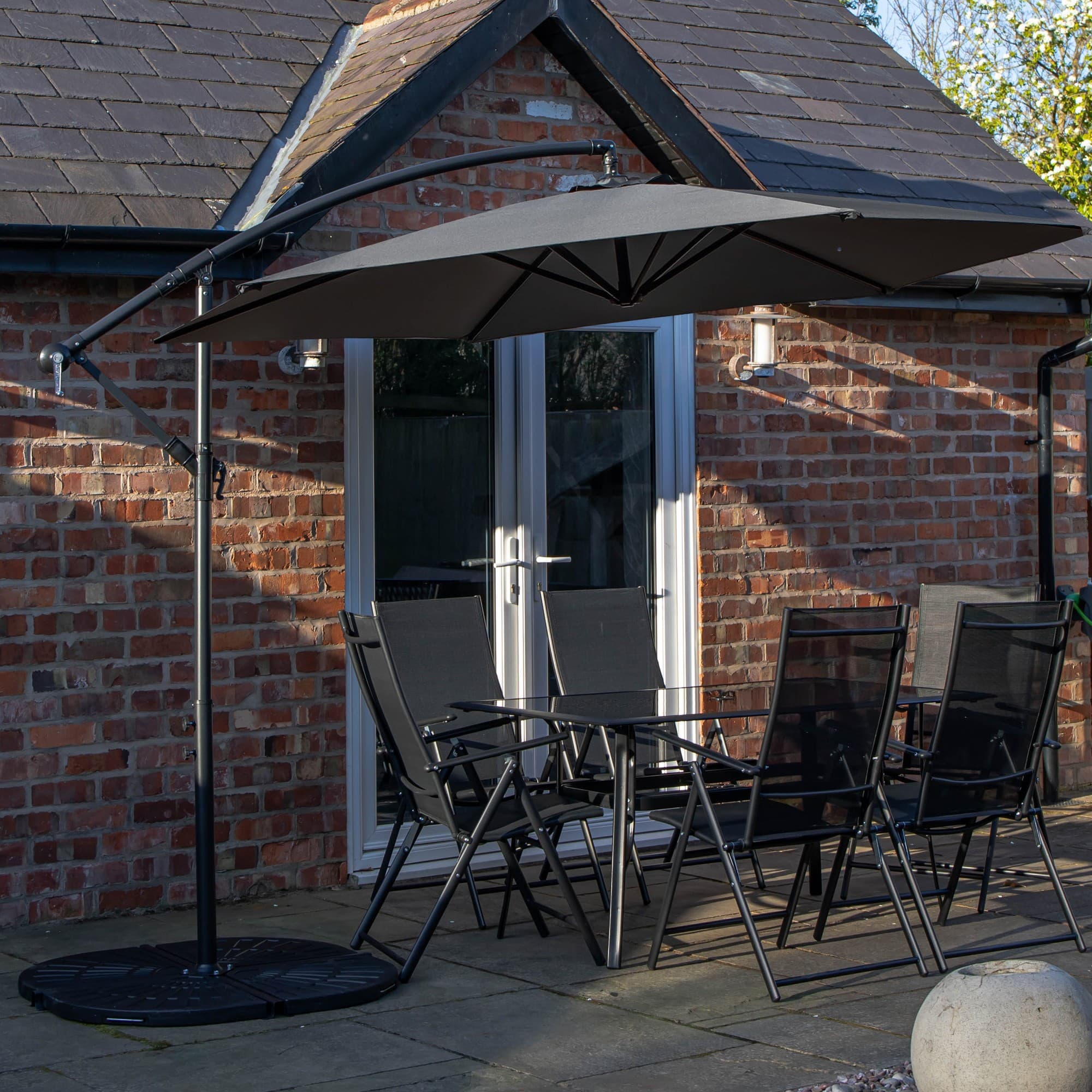 Rectangular glass top table surrounded by six black chairs, topped with a grey banana parasol and base, set on a patio with a brick wall backdrop.
